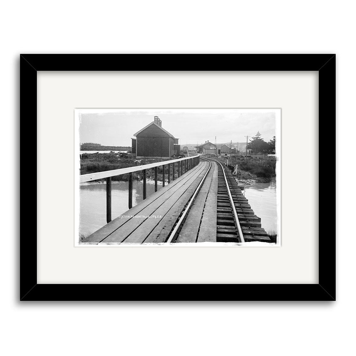Archival quality black and white photo titled Helensville Station 1914 - HI218, showing a single-track railroad curving over a raised wooden bridge toward a barn-like building, with water on both sides and utility poles in the background.
