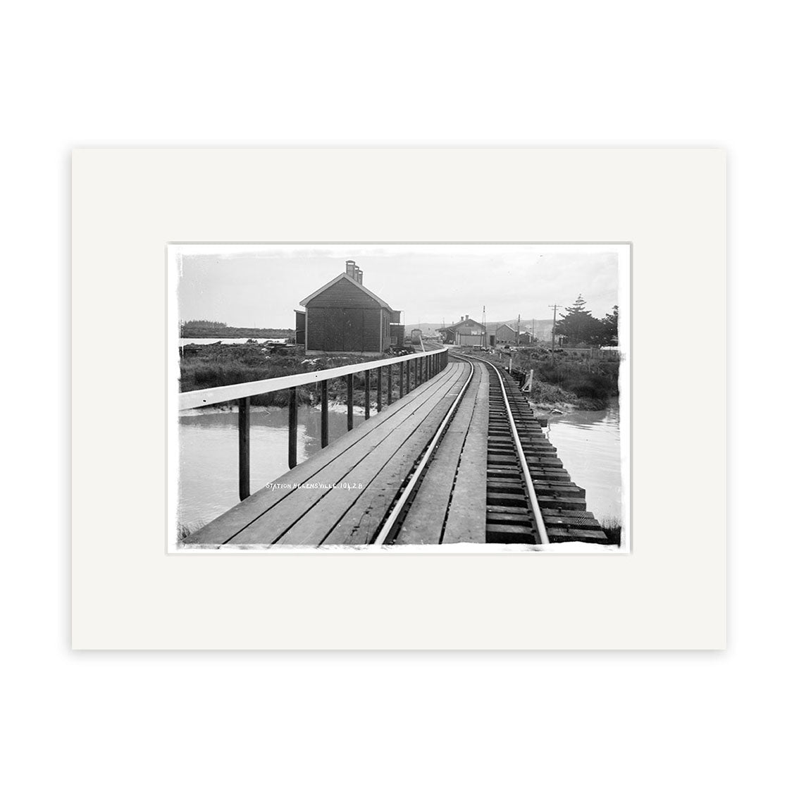 Archival quality historical photographic print of Helensville Station 1914 (HI218): black and white photo of train tracks and a wooden walkway by water in New Zealand, leading to a distant building with trees and utility poles.