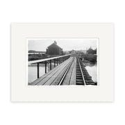 Archival quality historical photographic print of Helensville Station 1914 (HI218): black and white photo of train tracks and a wooden walkway by water in New Zealand, leading to a distant building with trees and utility poles.