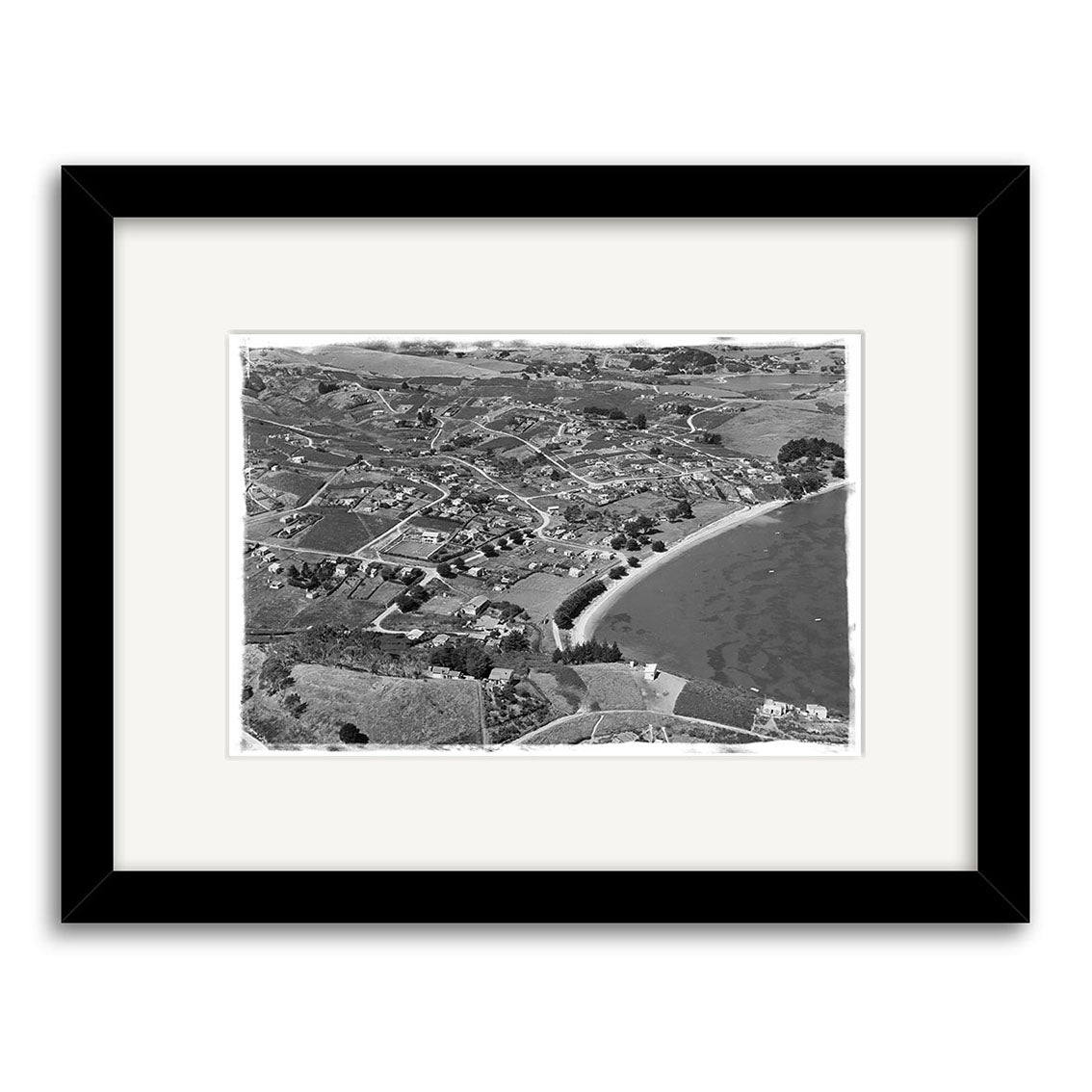 The Surfdale 1951 No.1 - HI219 is an archival quality black-and-white aerial photograph of a New Zealand coastal town, framed with a white mat and black frame. It shows scattered houses, roads, open fields, and a curved beach.