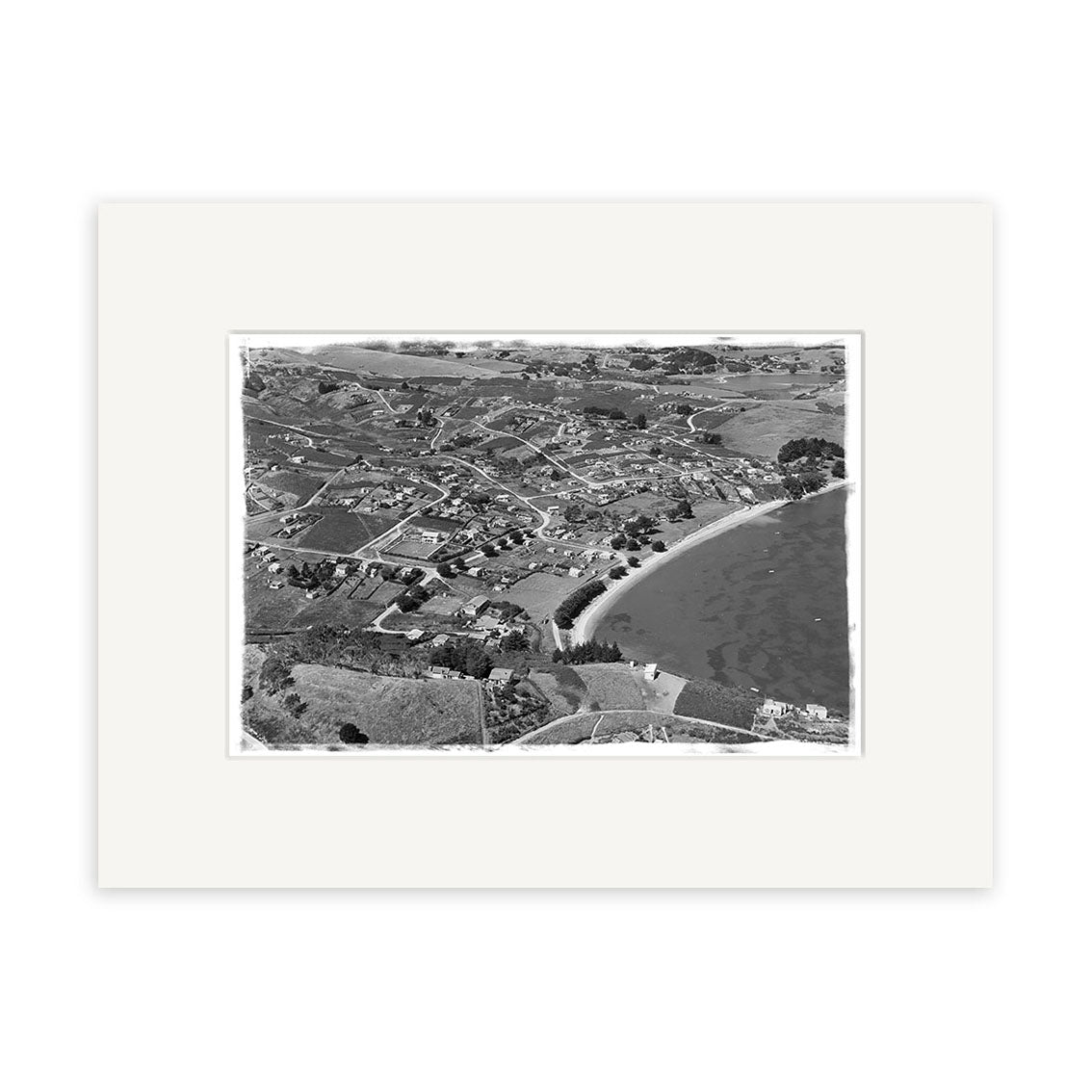 Surfdale 1951 No.1 - HI219 is a black and white aerial photograph of a New Zealand coastal town with houses, winding roads, and a curved sandy beach. The historical print is matted with a white border for archival display.