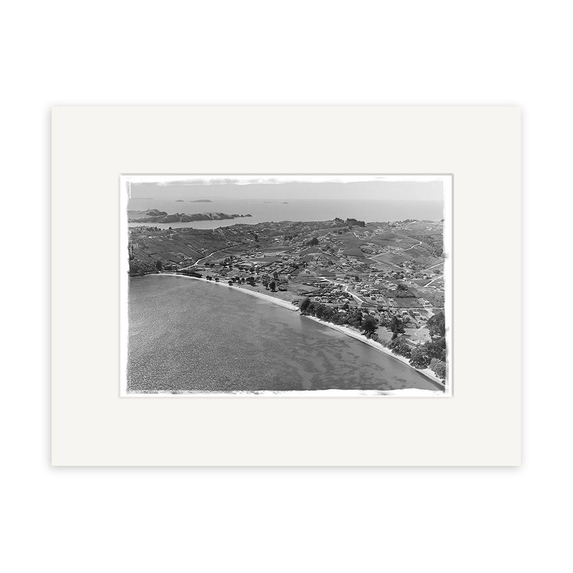 Surfdale 1951 No.2 - HI220 is a black-and-white aerial photo of a coastal New Zealand town with buildings, roads, farmland, and distant islands. This historical photographic print comes set within a white mat frame for archival quality.