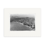 Surfdale 1951 No.2 - HI220 is a black-and-white aerial photo of a coastal New Zealand town with buildings, roads, farmland, and distant islands. This historical photographic print comes set within a white mat frame for archival quality.