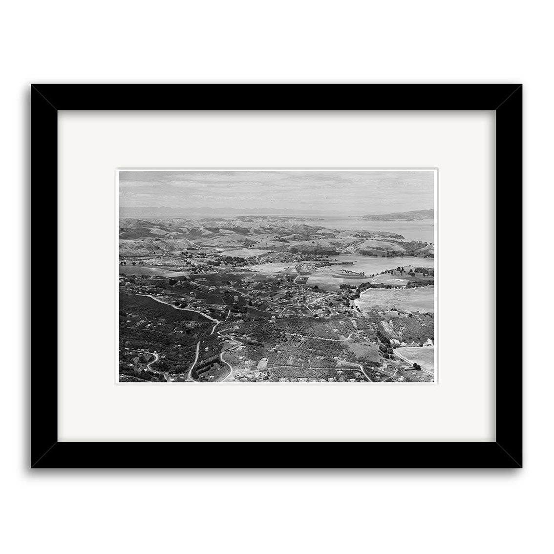 Archival-quality black-and-white aerial photo of a New Zealand coastal landscape—“Surfdale 1958 - HI221”—showing hills, roads, and buildings. Framed with a wide white mat and simple black frame.