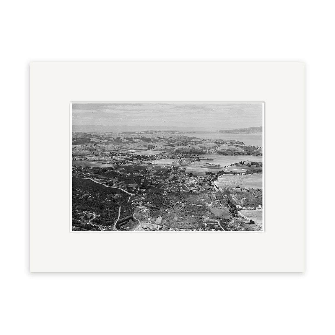 The black-and-white aerial photograph Surfdale 1958 - HI221 captures coastal New Zealand with hills, roads, buildings, and water in the distance. Displayed in an archival quality white mat board.