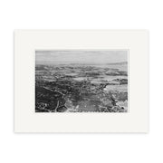 The black-and-white aerial photograph Surfdale 1958 - HI221 captures coastal New Zealand with hills, roads, buildings, and water in the distance. Displayed in an archival quality white mat board.