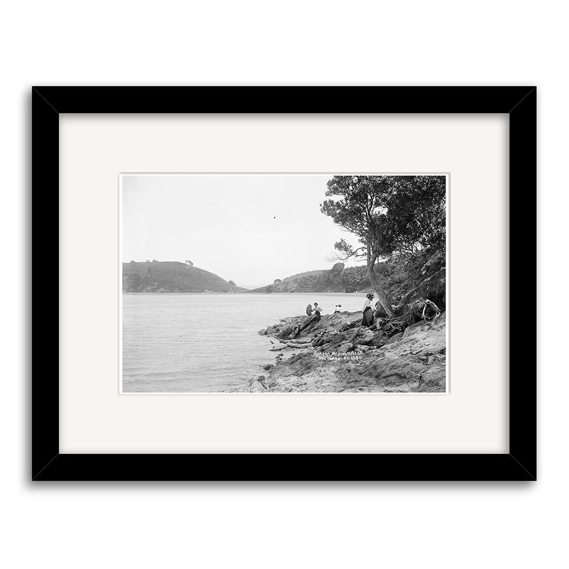 Archival quality black and white photo print of people on the rocky shoreline of Awaroa Bay, New Zealand, 1920 (HI222), with calm water, hills, and trees; presented with a white mat and black frame.
