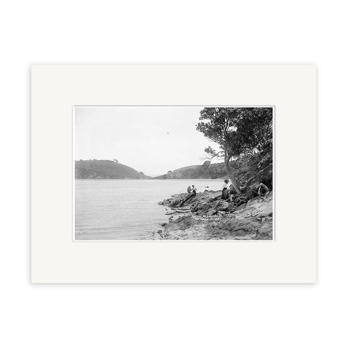 Archival-quality black and white photographic print, Awaroa Bay 1920 - HI222: features New Zealand shoreline, people on rocky ground near water, trees on the right, and hills in the background—a glimpse into the past.