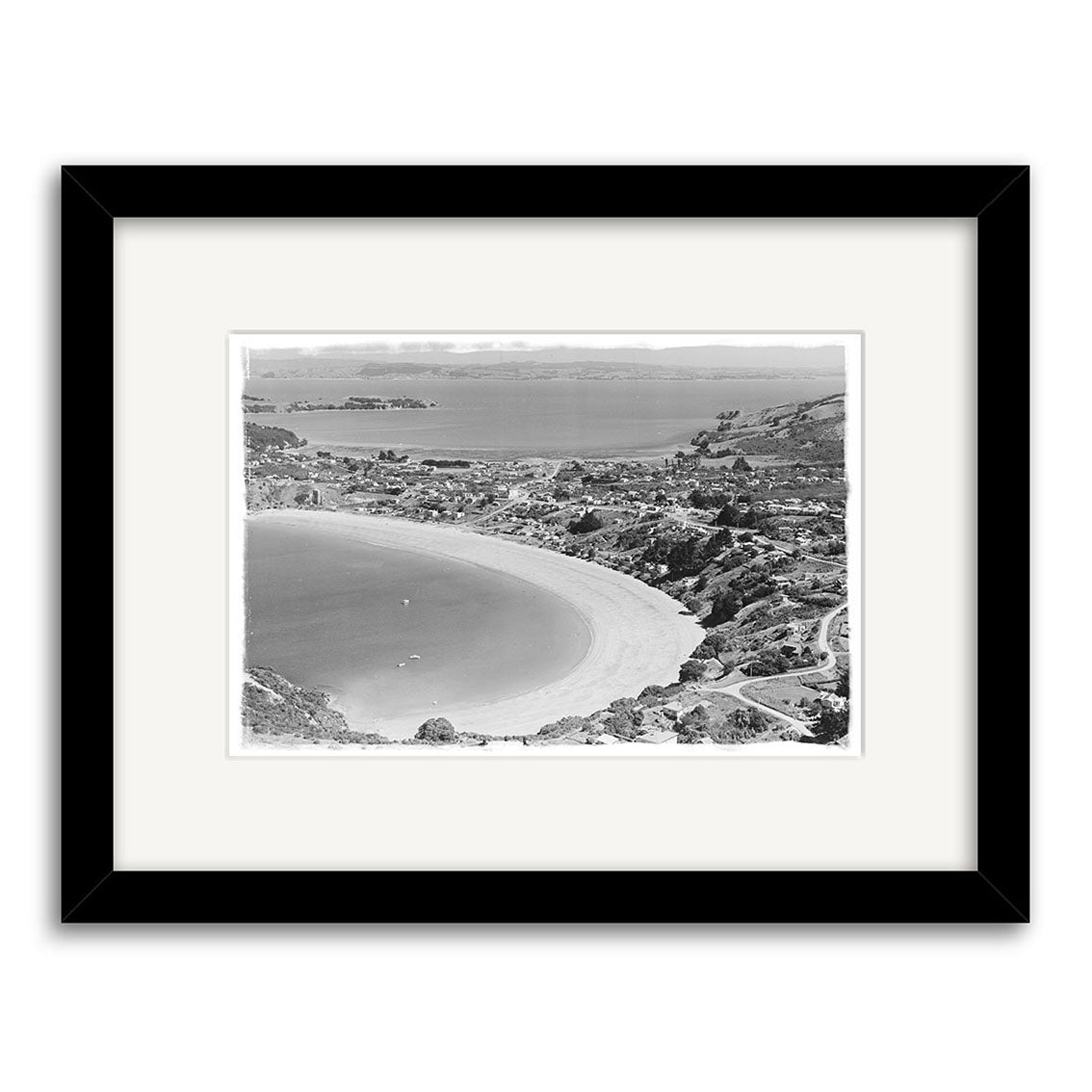 Oneroa Waiheke 1961 - HI223: Black-and-white historical aerial photo of a curved beach and coastal town in New Zealand, on archival quality paper, framed with a white mat and black frame.