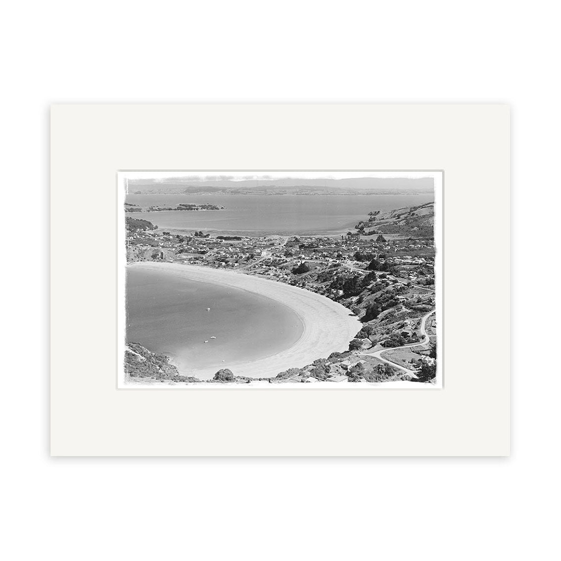 Oneroa Waiheke 1961 - HI223: Black and white aerial photo of a New Zealand coastal town with a curved beach, hills, houses, and roads. Printed on archival paper with a white mat.