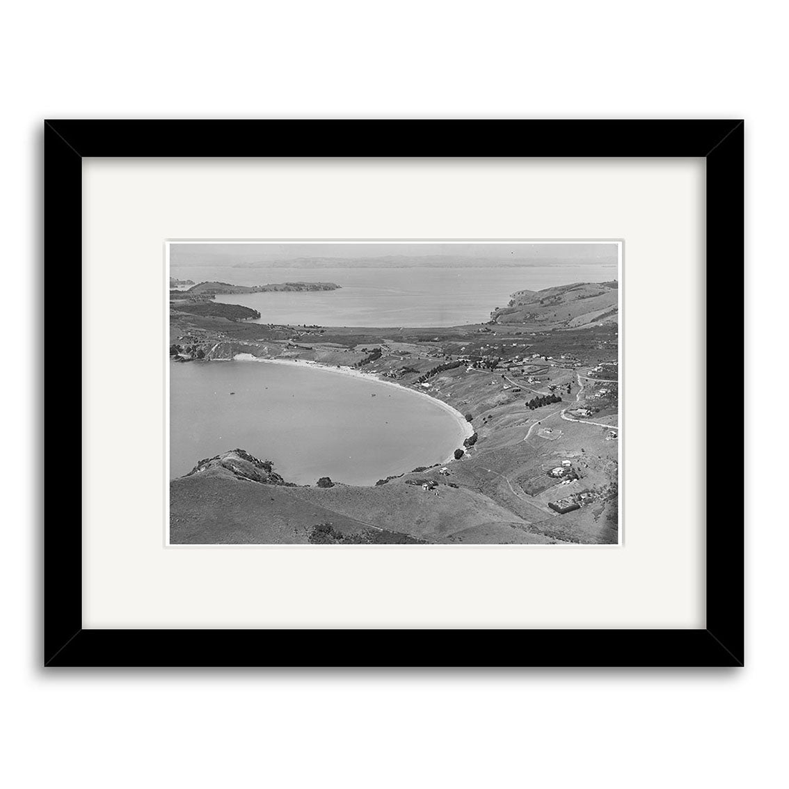 Black-and-white historical photograph titled Oneroa Waiheke 1938 - HI224 shows a curved shoreline, hills, scattered buildings, and water. Presented in a black frame with white mat on archival matte paper.