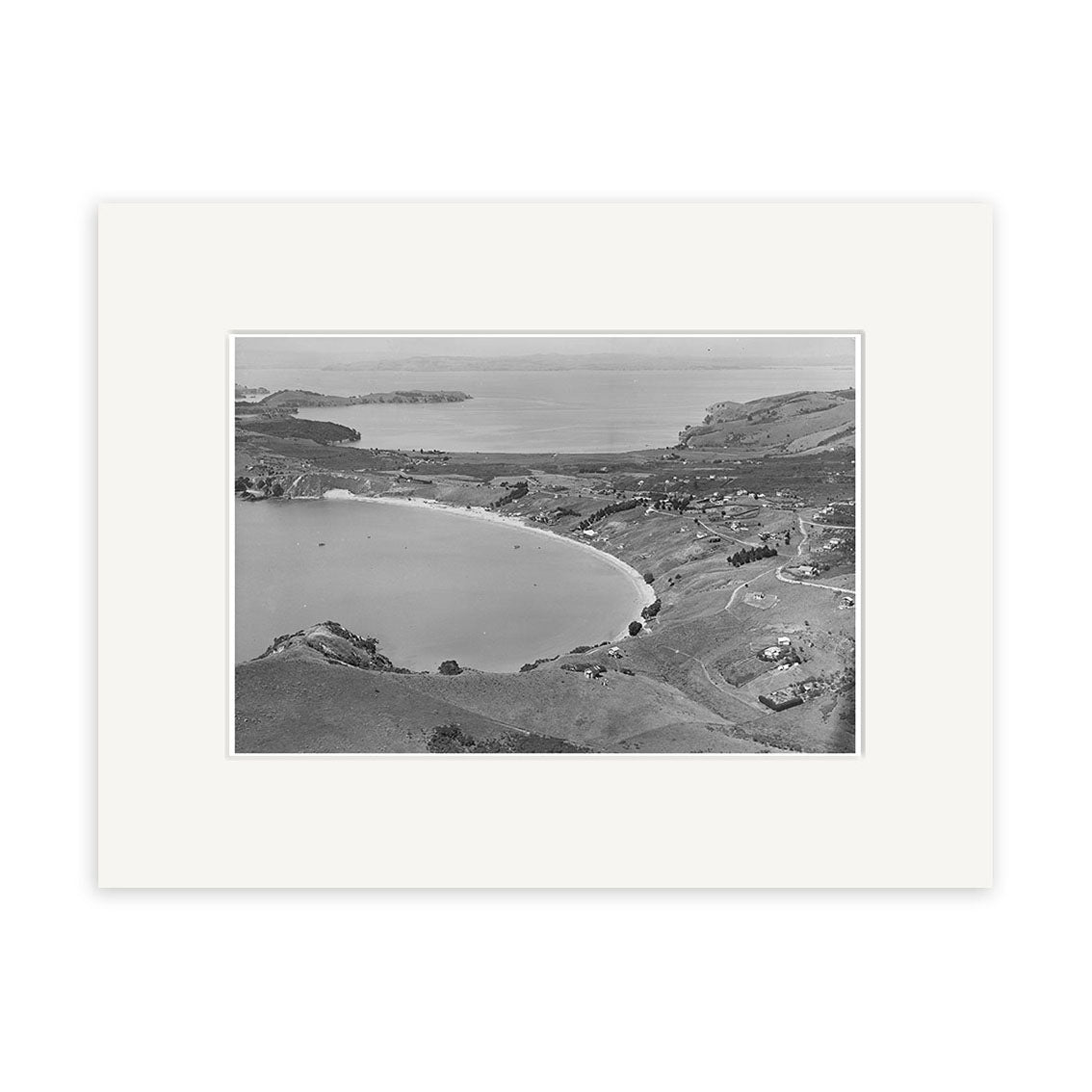 Oneroa Waiheke 1938 - HI224: A black and white aerial photo on archival matte paper captures a curved bay, gentle hills, scattered buildings, open land, and distant water stretching to the horizon.