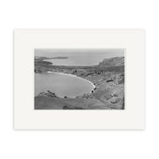 Oneroa Waiheke 1938 - HI224: A black and white aerial photo on archival matte paper captures a curved bay, gentle hills, scattered buildings, open land, and distant water stretching to the horizon.