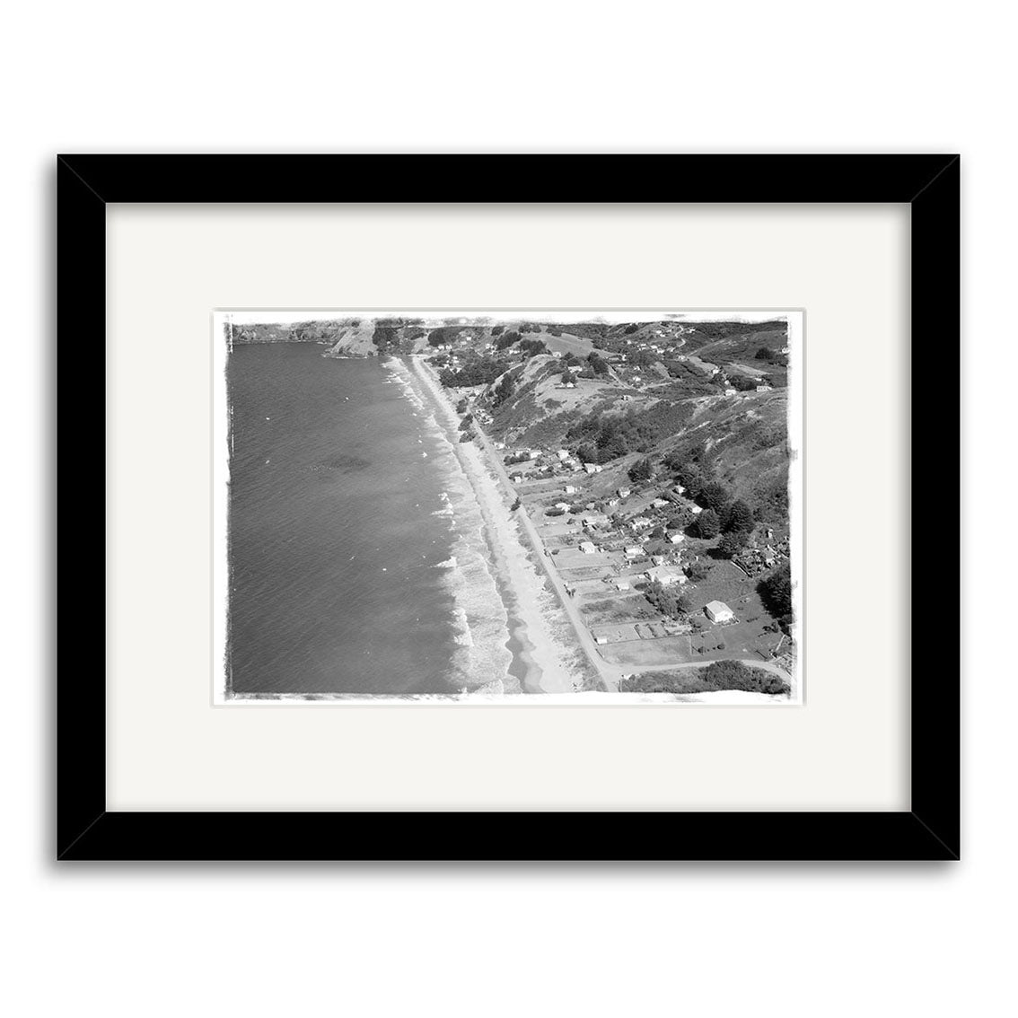 The Onetangi 1963 - HI225 is a framed archival matte print of a black and white aerial photo depicting a New Zealand coastal town with shorefront houses, waves on the sandy beach, and tree-covered hills. Features white mat and black frame.