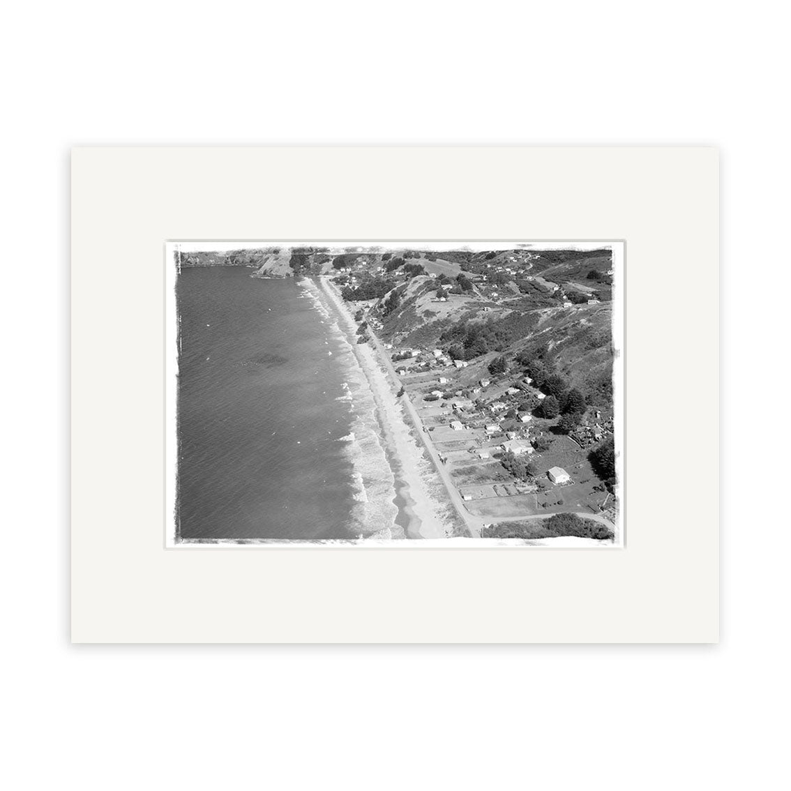 The historical photographic print Onetangi 1963 - HI225 features a black and white aerial view of a New Zealand coast, with waves, beachfront houses, and tree-covered hills, displayed with a white mat border.