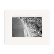 The historical photographic print Onetangi 1963 - HI225 features a black and white aerial view of a New Zealand coast, with waves, beachfront houses, and tree-covered hills, displayed with a white mat border.