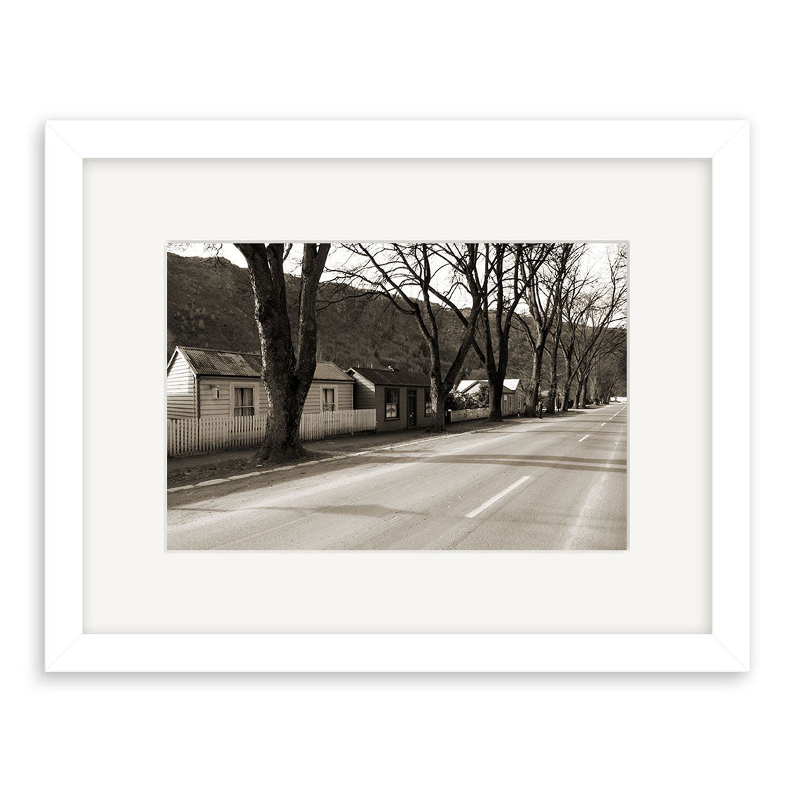 Arrowtown - RP068 is a black-and-white photographic print of a quiet New Zealand street with bare trees and small houses, framed in white. The empty road leads to distant hills, captured in archival quality.