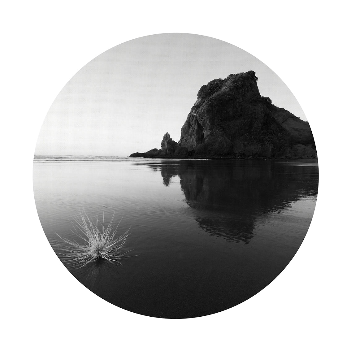 Piha Art Spot features a black and white beach scene with a large rock reflected in wet sand and grass in the foreground, framed in a circle. Printed on matte fabric, this repositionable wall art is perfect for easy decor updates.