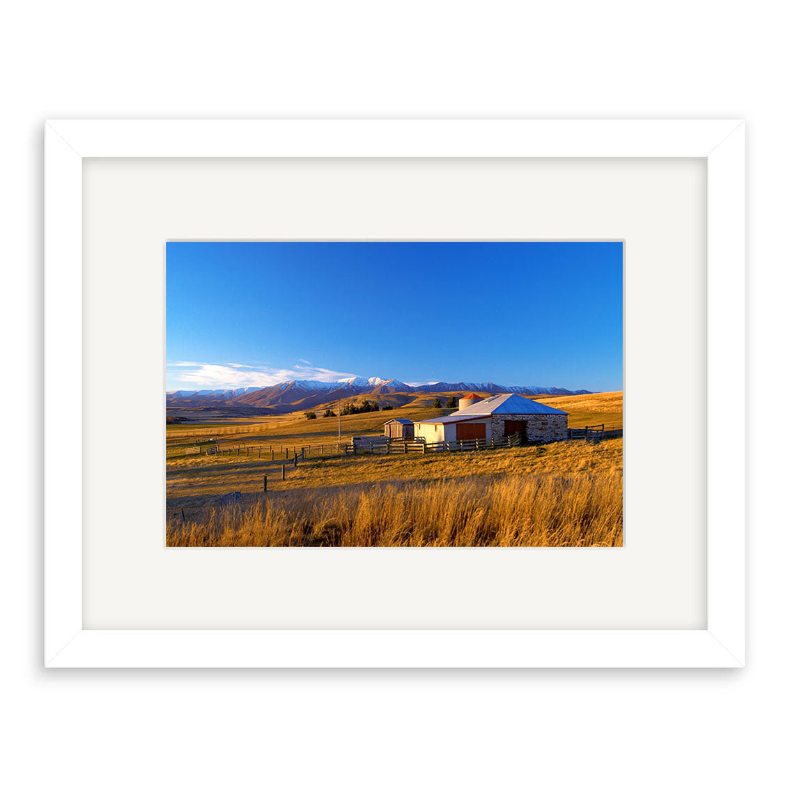 Central Otago - RP074 is a framed photographic print by Reuben Price, depicting a rural New Zealand landscape with a small house, barn, golden fields, and snow-capped mountains under a clear blue sky.