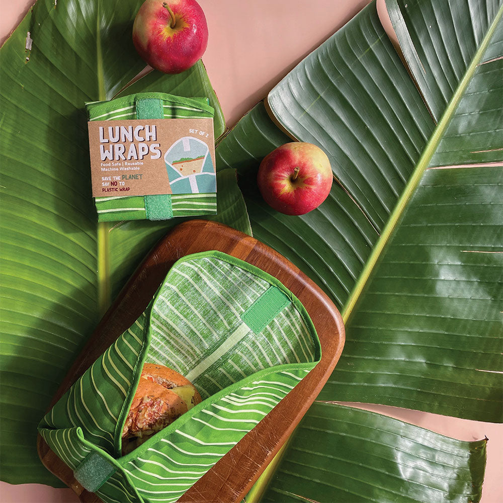 A sandwich in a green Pop Art Banana Lunch Wrap sits on a wooden board with two red apples, its packaging, and large banana leaves on a pink surface. The plastic-free wrap is food-safe certified for eco-friendly lunches.
