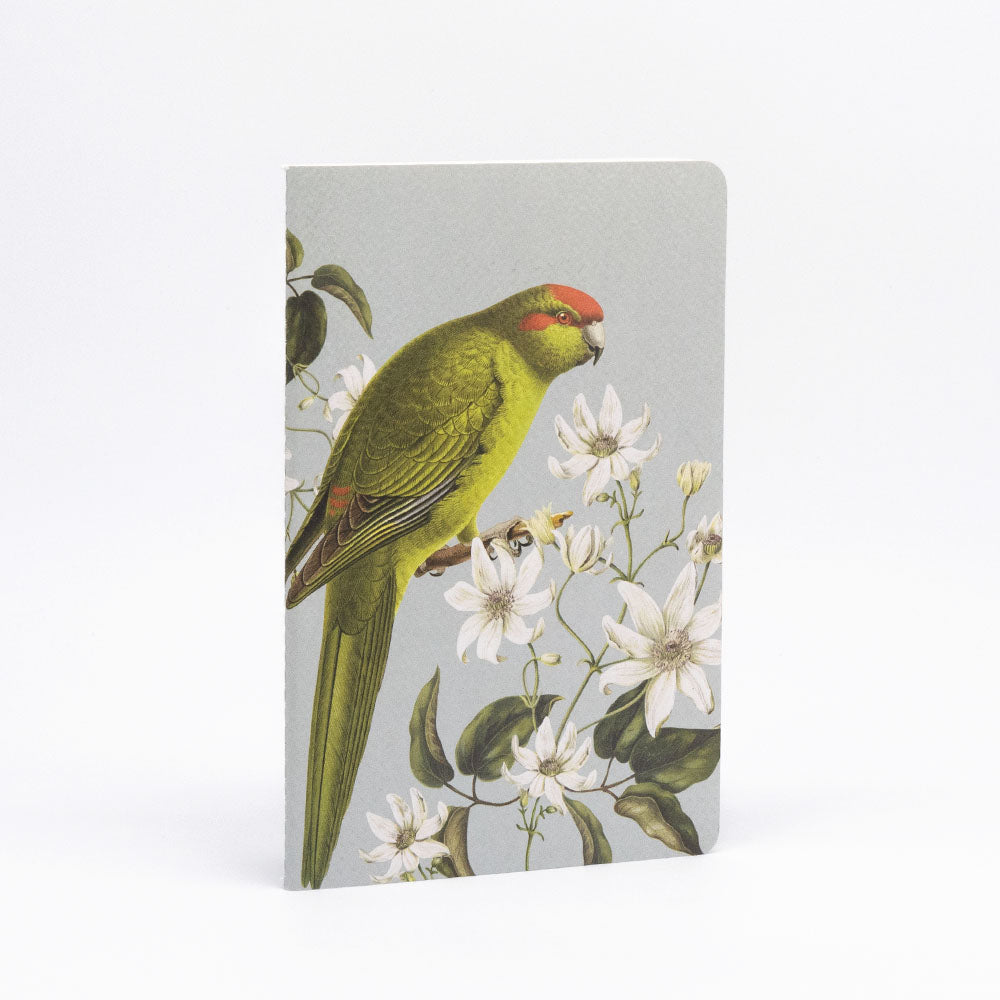 The Birds & Botanicals Notebook Set features a lined notebook with a green parrot on a branch with white flowers against a light gray cover, hand-stitched binding, and quality paper for an enjoyable writing experience.