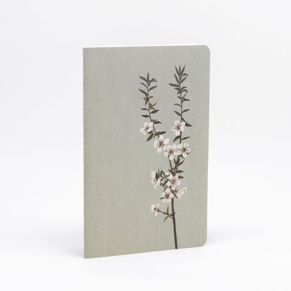 The Vintage Botanicals Notebook Set includes a hand-stitched notebook with a light grey cover, decorated with an illustrated branch of white flowers and green leaves, shown standing upright against a plain white background.