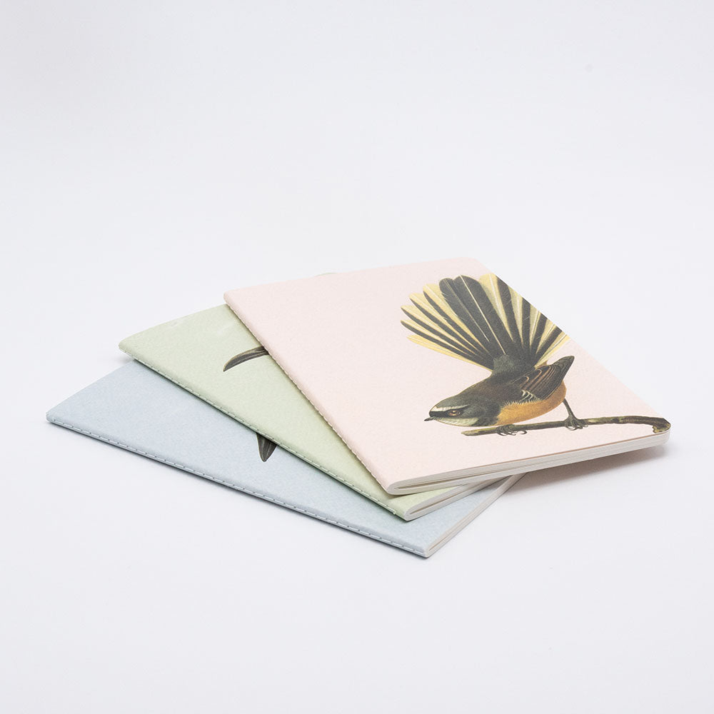 The Hushed Birds Notebook Set features three hand-stitched notebooks with pastel covers, stacked on a white surface. The top notebook sports a beige cover adorned with an illustration of a bird perched on a branch.