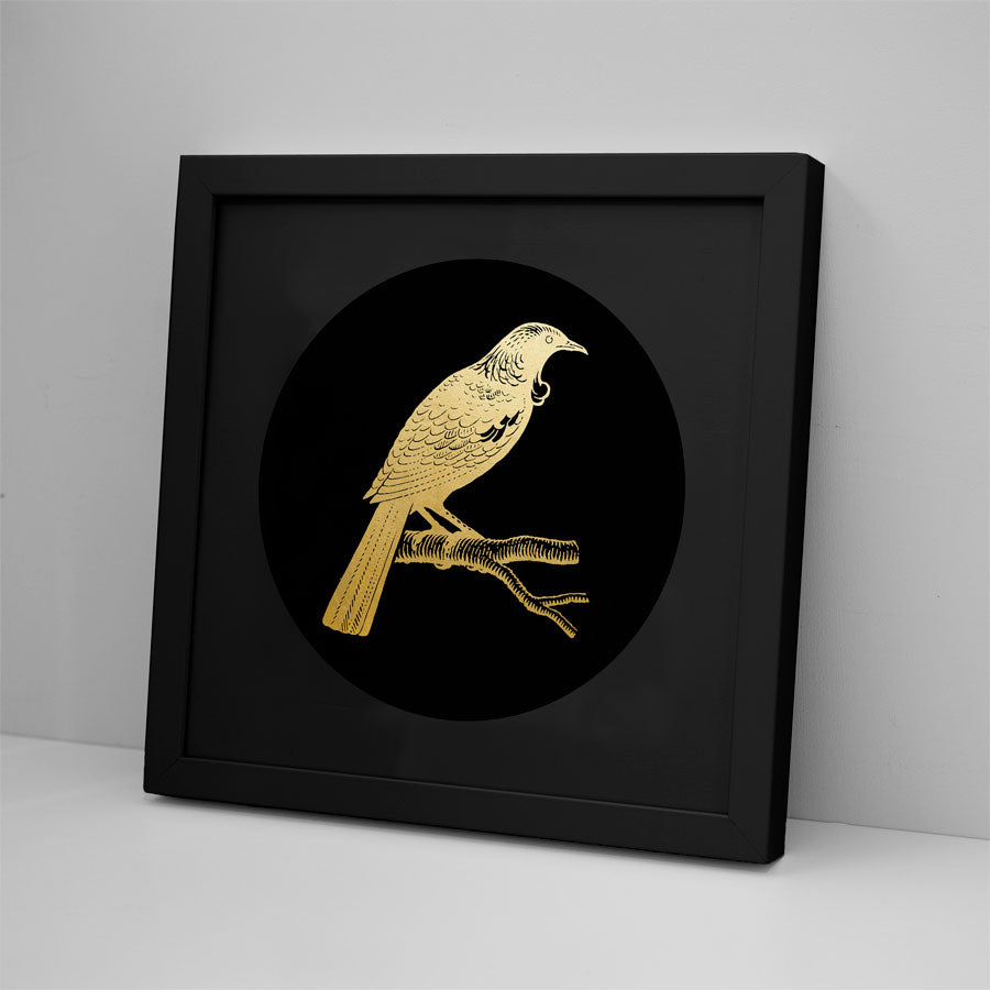 The Tui Foil Print is a framed gold foil artwork of a bird perched on a branch, set against a black circular background. Its black-on-black design and sleek frame add elegance when displayed on a white surface near a plain white wall.
