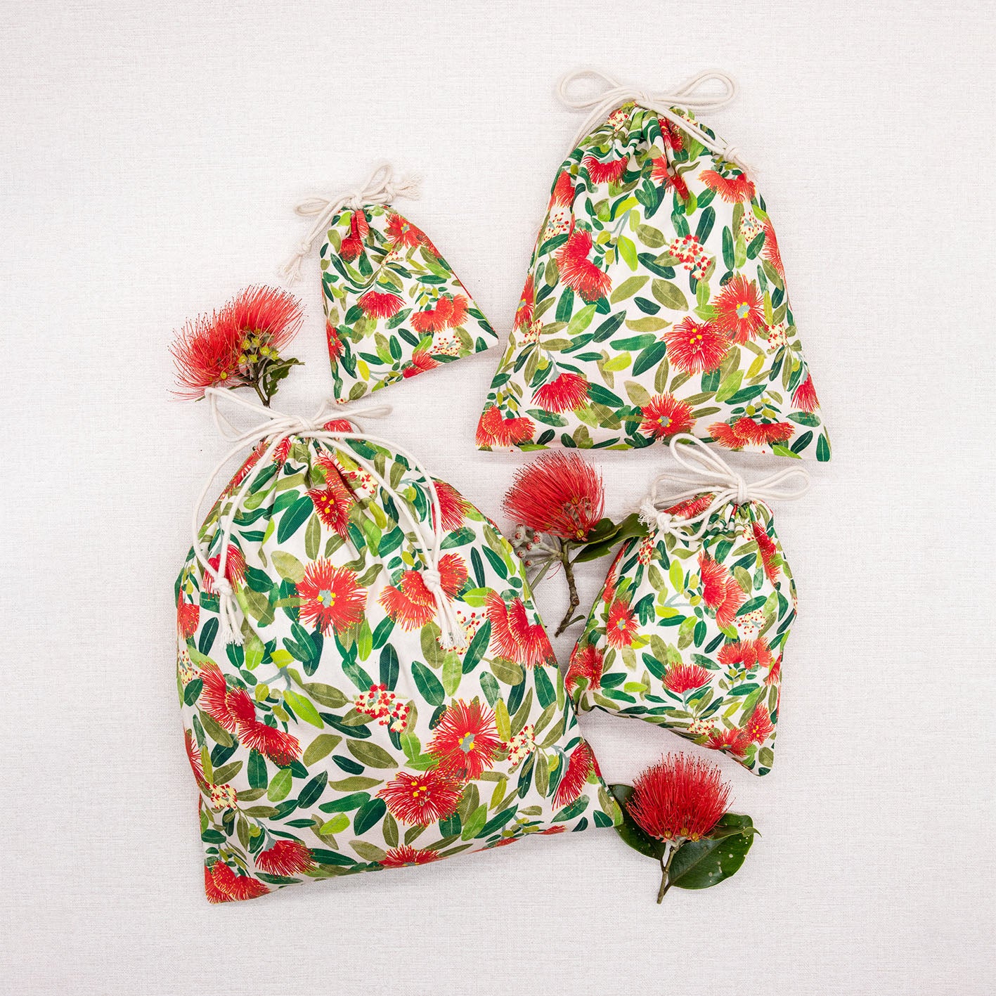 The Pohutukawa Print Drawstring Bag Set includes four cotton storage bags in various sizes with vibrant prints, shown on a white surface beside red flowers and green leaves—perfect for festive, reusable organization.