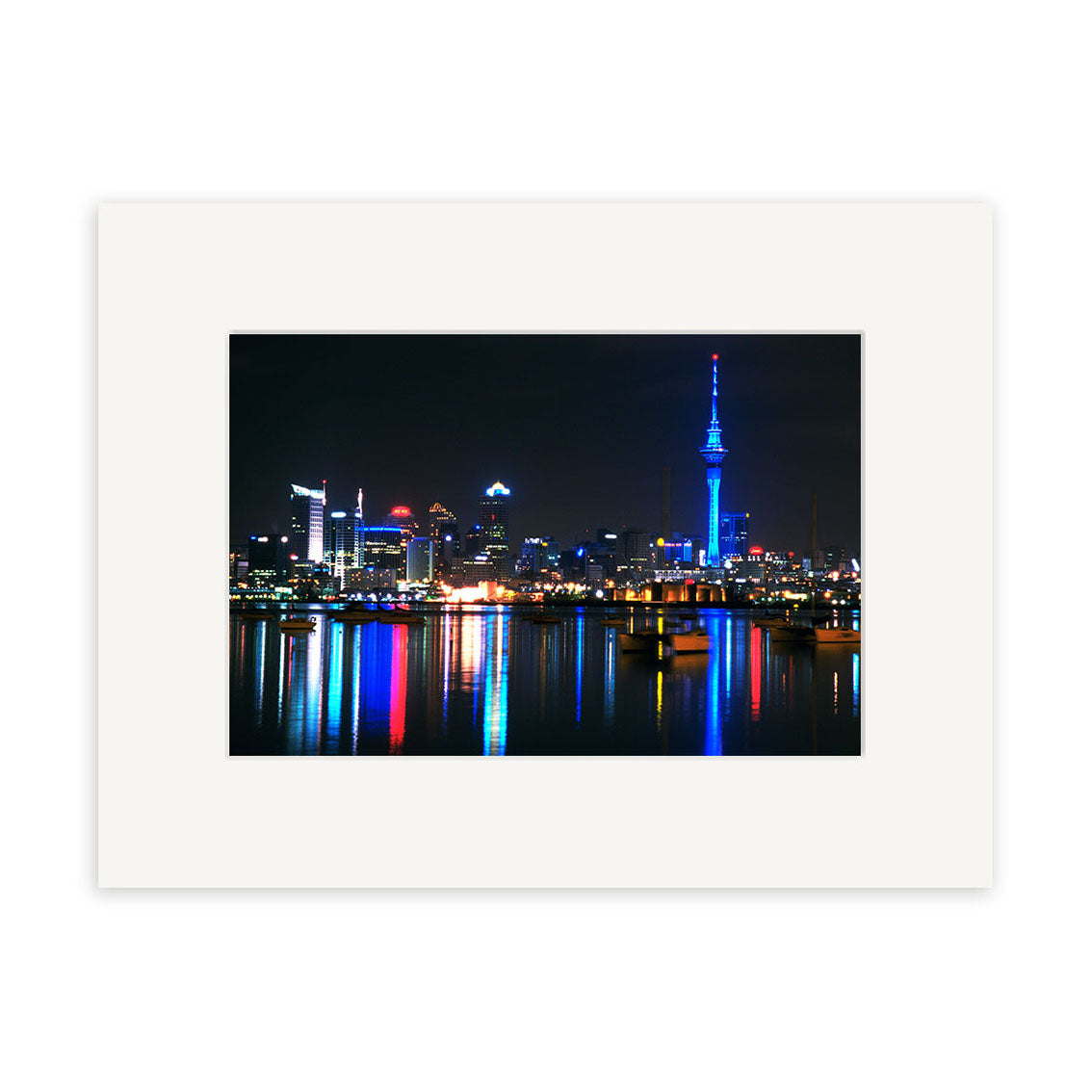 Auckland City by Night - RP007 showcases illuminated buildings and a blue-lit tower reflecting vibrant lights on the water. Printed on archival matte paper with a white border, its an ideal decor piece for New Zealand enthusiasts.