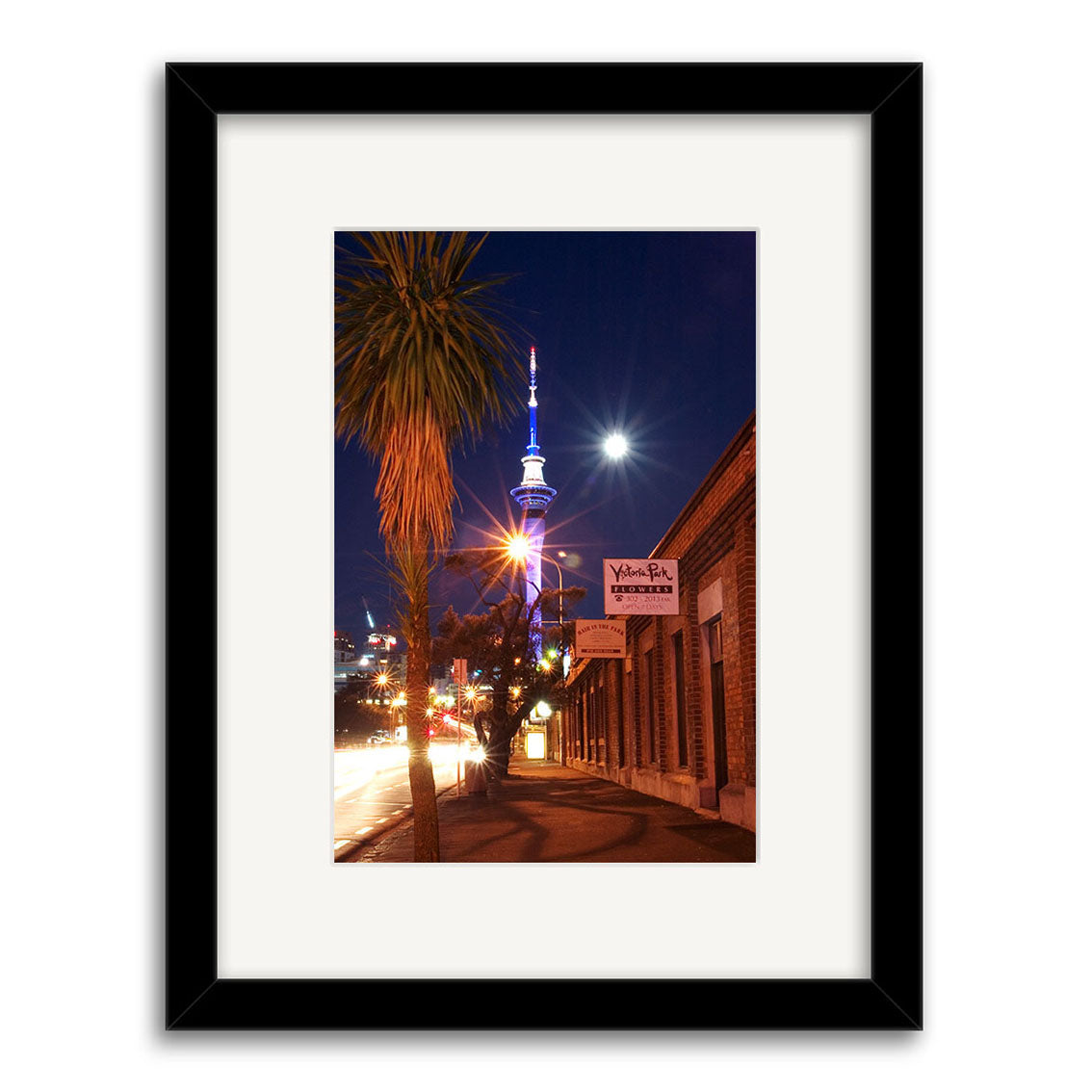 The Victoria Park - RP008 is a framed photo of a New Zealand city street at night with palm trees, brick buildings, and the Sky Tower beneath a bright moon, printed on archival-quality matte paper for lasting brilliance.