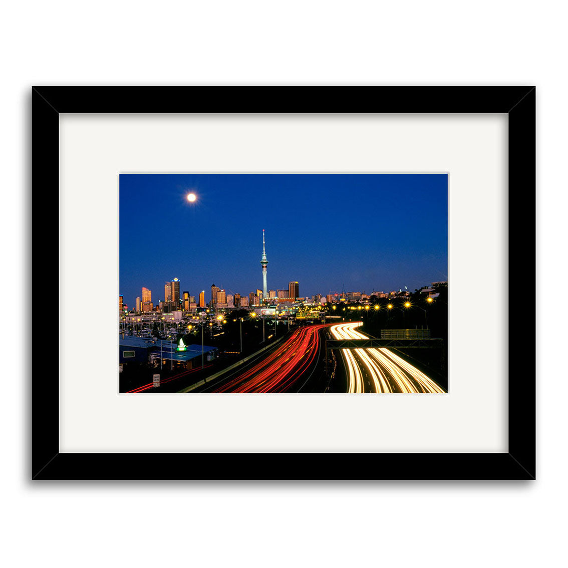 Twilight Cars - RP010 is a framed photographic print of a city skyline at night with illuminated buildings, a central tower, a bright moon, and highway car light trails—archivally crafted for lasting beauty.