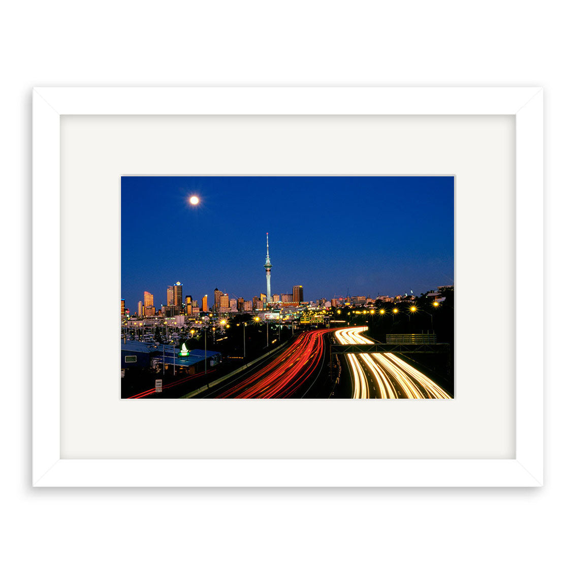 Twilight Cars - RP010: Framed photographic print featuring a cityscape at dusk with light trails, a central tower, buildings, and a full moon—crafted with archival quality for lasting beauty.