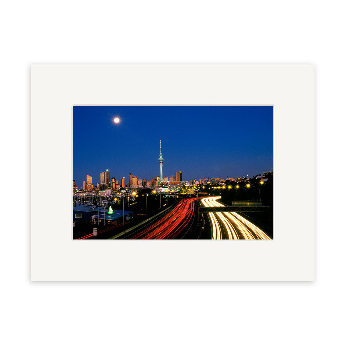 Twilight Cars - RP010: A framed cityscape print of New Zealand at night, showcasing a brightly lit tower, city buildings, full moon, and car light trails—crafted with archival quality for lasting beauty.