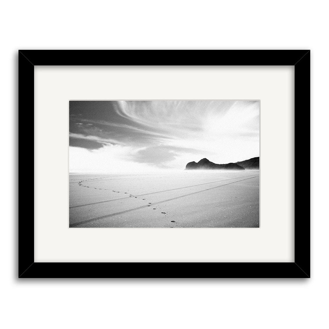 Bethells Beach - RP015: Black-and-white photo of a New Zealand beach with footprints in the sand, distant rocky hills, and wispy clouds. Archival matte print, white mat, black frame.