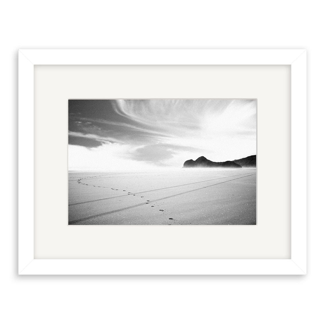 Bethells Beach - RP015: Black and white photo of a sandy New Zealand beach with footprints curving toward distant hills under a cloudy sky, framed with a white mat and frame. Available as archival quality matte photographic prints.