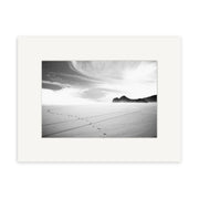 Bethells Beach - RP015: A black and white photo of footprints on a smooth sandy New Zealand beach with distant rocky hills and clouds, bordered by a wide white mat on archival quality matte paper.