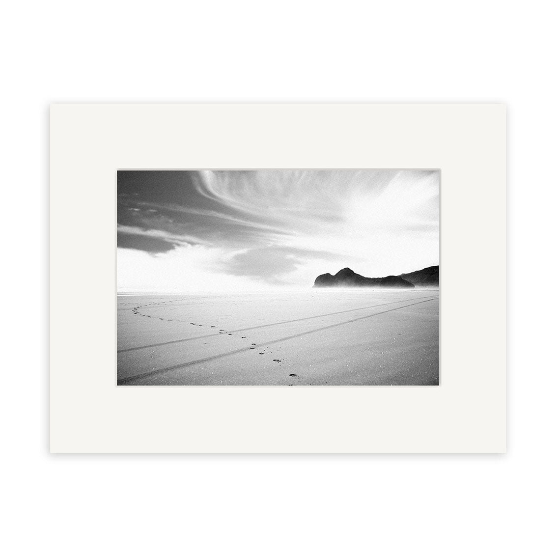 Bethells Beach - RP015: A black and white photo of footprints on a smooth sandy New Zealand beach with distant rocky hills and clouds, bordered by a wide white mat on archival quality matte paper.