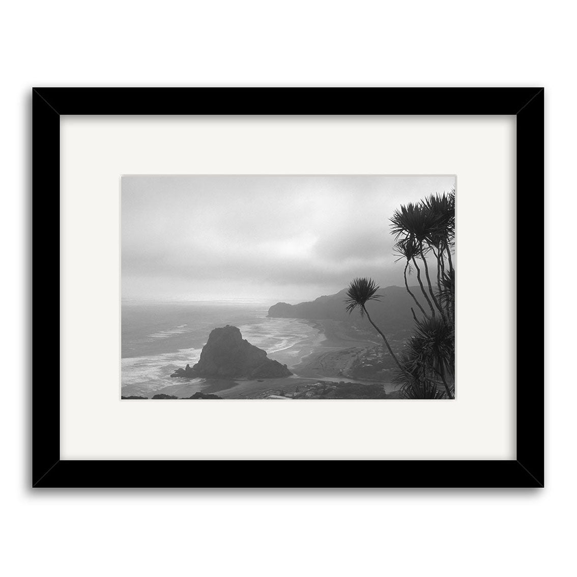 Black and white photo Piha Beach - RP016 by Reuben Price features a rocky New Zealand coastline with waves, cliffs, and spiky plants. Presented in a black frame with white mat, printed on archival matte paper.
