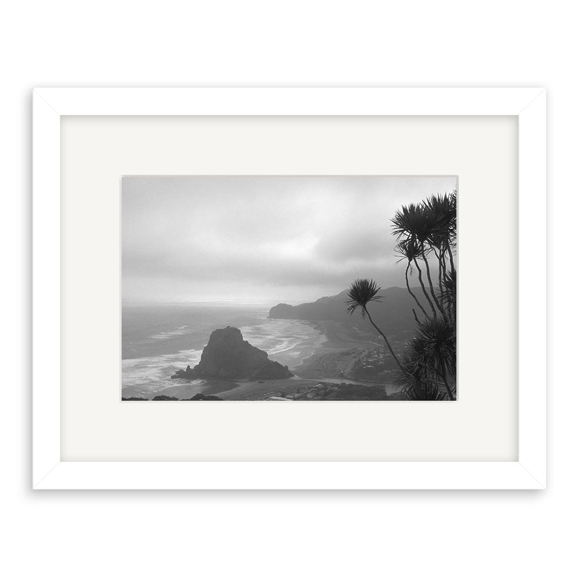 Black and white photograph of Piha Beach (RP016) by Reuben Price, featuring a coastal landscape with a large rock formation, waves, and spiky plants. Presented in a white mat and frame, printed on archival quality matte paper.