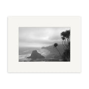 Black and white photo Piha Beach - RP016 by Reuben Price features waves, a large rock, spiky plants, and a cloudy sky. Printed on archival quality matte paper and framed with a white mat.