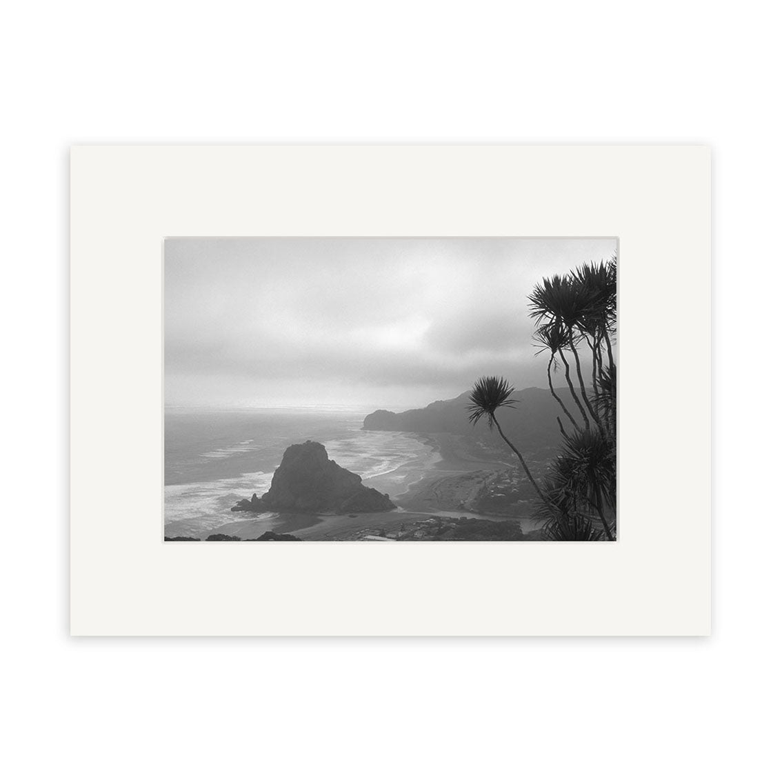 Black and white photo Piha Beach - RP016 by Reuben Price features waves, a large rock, spiky plants, and a cloudy sky. Printed on archival quality matte paper and framed with a white mat.
