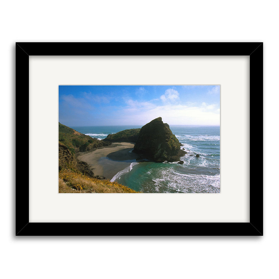 Framed photographic print, Piha South - RP017, features a rocky New Zealand coast with a large rock formation, sandy beach, waves, and blue sky. Presented with a white mat and black frame on archival matte paper.