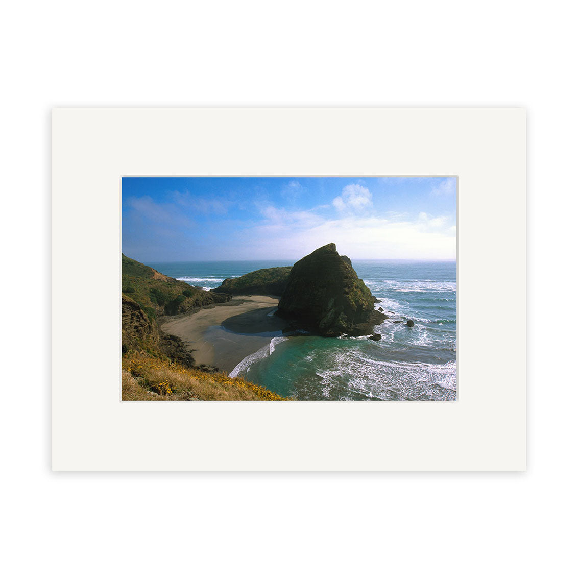 Piha South - RP017 captures a rocky coastal outcrop, ocean waves, sandy beach, grassy cliffs, and a blue sky with light clouds in a stunning photographic print on archival quality matte paper.