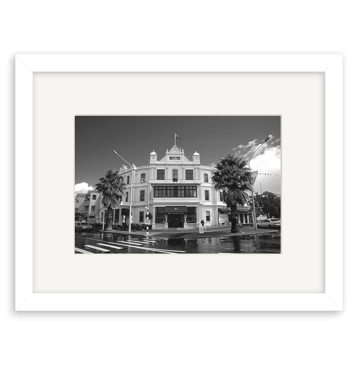 Devonport Esplanade - RP023: Black and white photo of a historic multi-story New Zealand building with balcony, columns, and flag, framed by palm trees from a street corner, displayed in a white mat on archival quality paper.