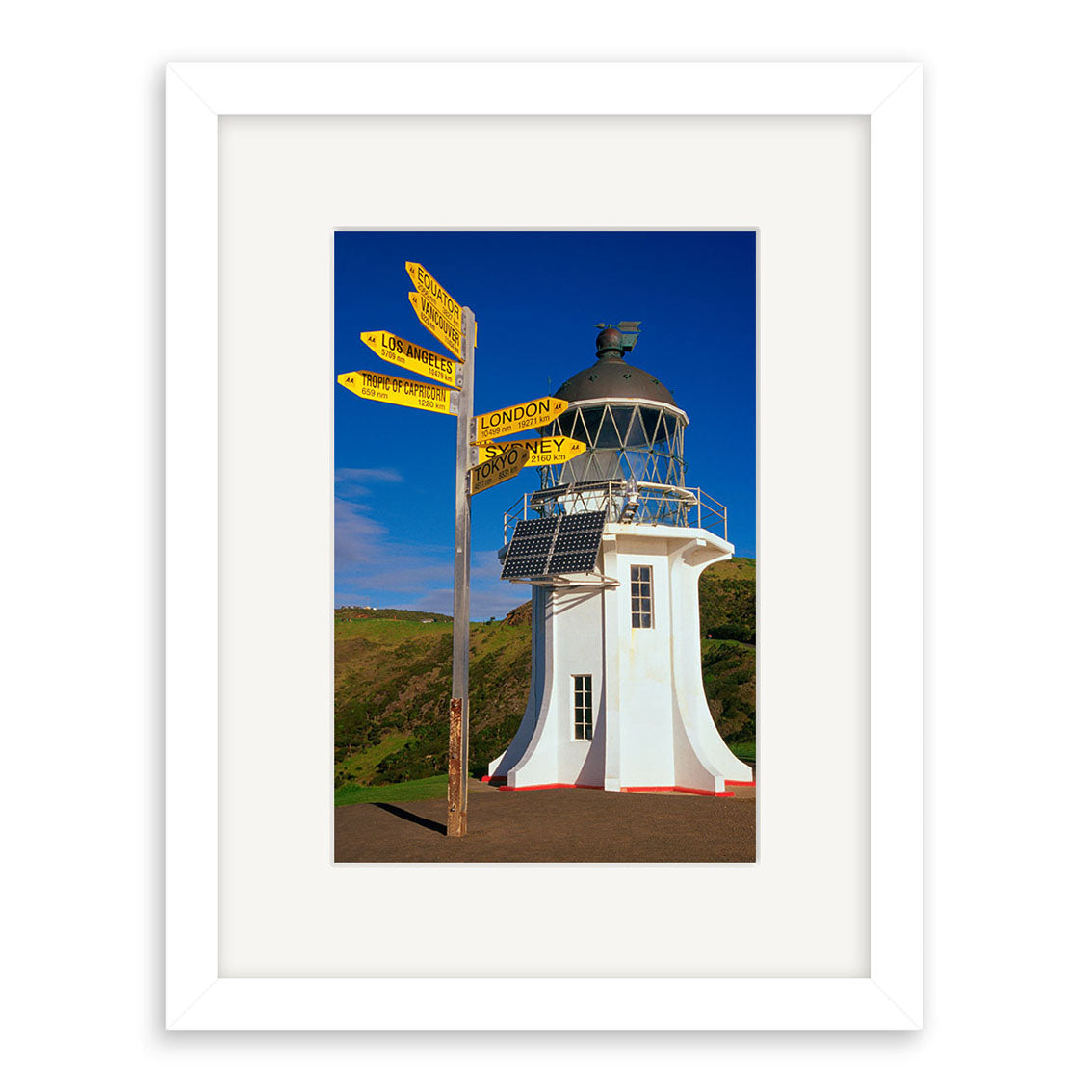 The Cape Reinga Lighthouse - RP024 features a white lighthouse in New Zealand, yellow city signposts, blue sky, and green hills. Its printed on archival matte paper and presented in a white frame with mat.