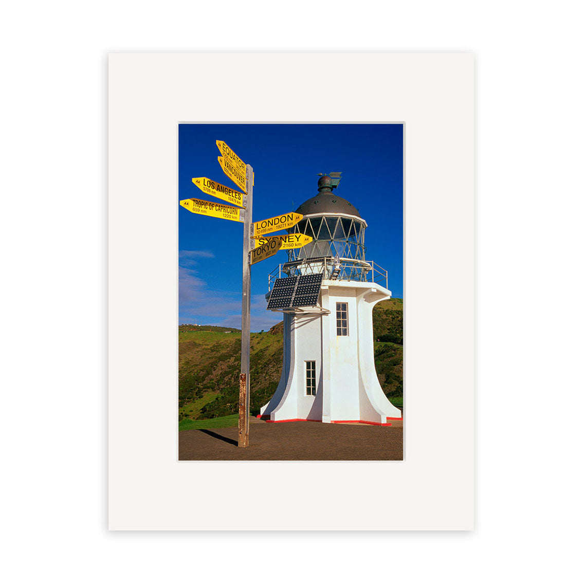 Cape Reinga Lighthouse - RP024 features a white lighthouse with a glass dome beside a wooden post of yellow signs to London, Sydney, and NZ cities, set against blue sky and green hills. Ideal for photographic prints on archival matte paper.