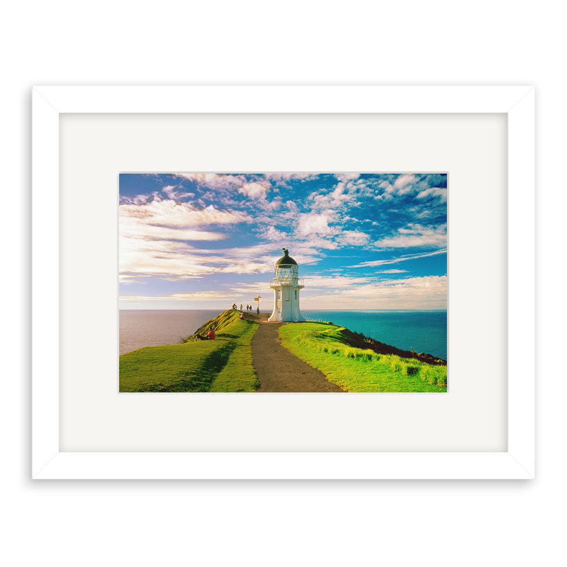 Cape Reinga - RP025: A framed photographic print of a white lighthouse on a grassy New Zealand cliff overlooking the ocean, under a clear sky with scattered clouds. A paved path leads to the lighthouse. Printed on archival quality paper.