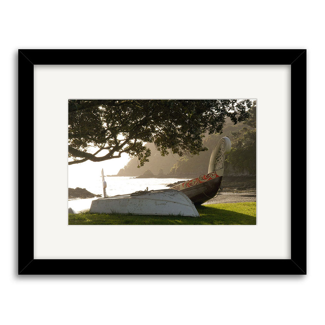 Waka - RP027: A framed photographic print on archival quality matte paper, featuring two boats on grass by a New Zealand shoreline—one a carved wooden canoe in shadow under a tree with coastal water and distant hills.