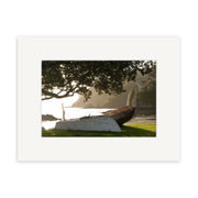The Waka - RP027, a carved wooden canoe with decorative patterns, rests on grass near a New Zealand shoreline, partially shaded by a tree, with water and rocky coastline in the background.